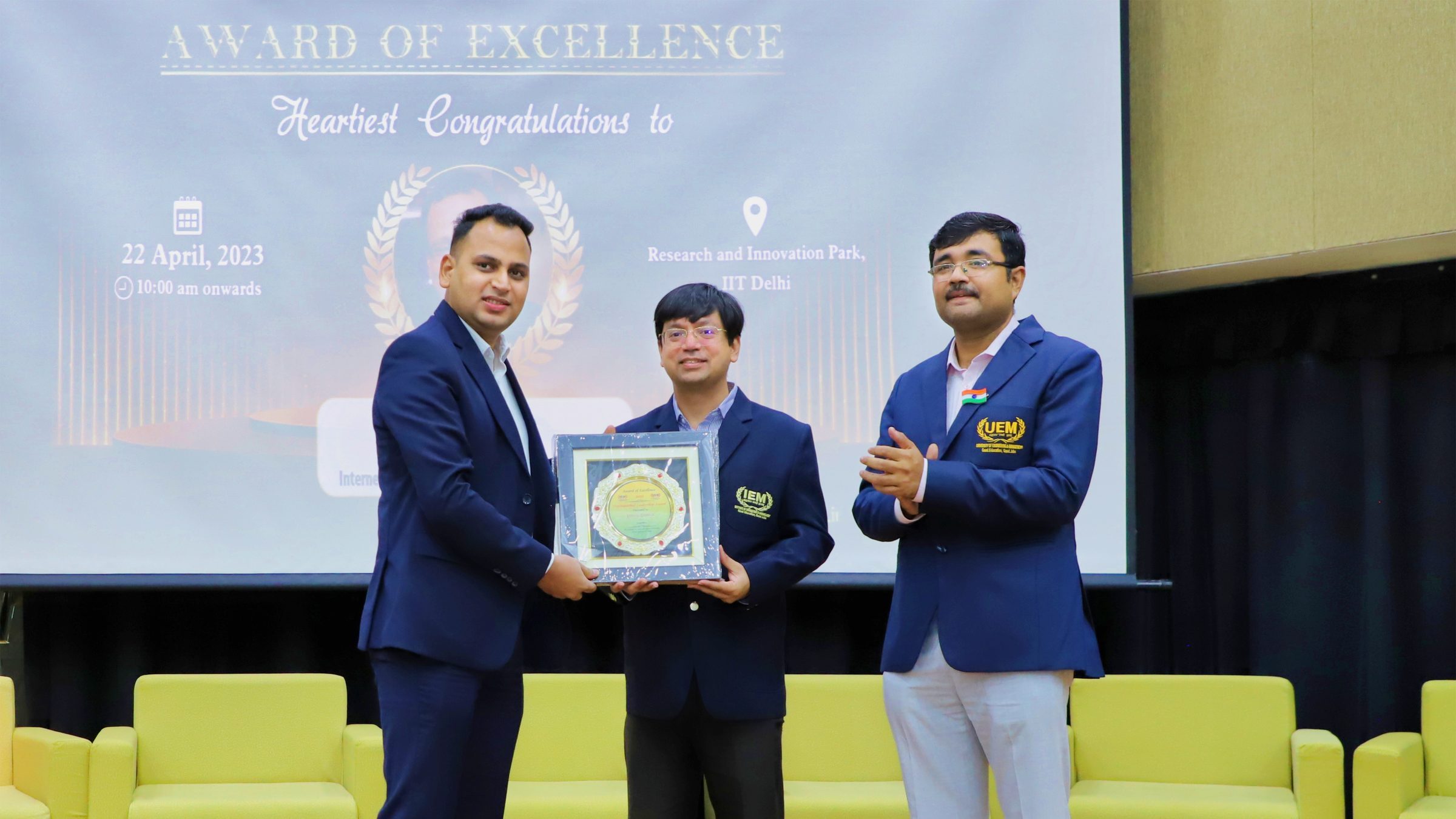 Lithvik Mukesh Sharma stands on stage at IIT Delhi receiving the Award of Excellence, presented by representatives of the University of Engineering and Management. The large projection screen behind them displays the Award of Excellence alongside a circular laurel-wreath graphic
