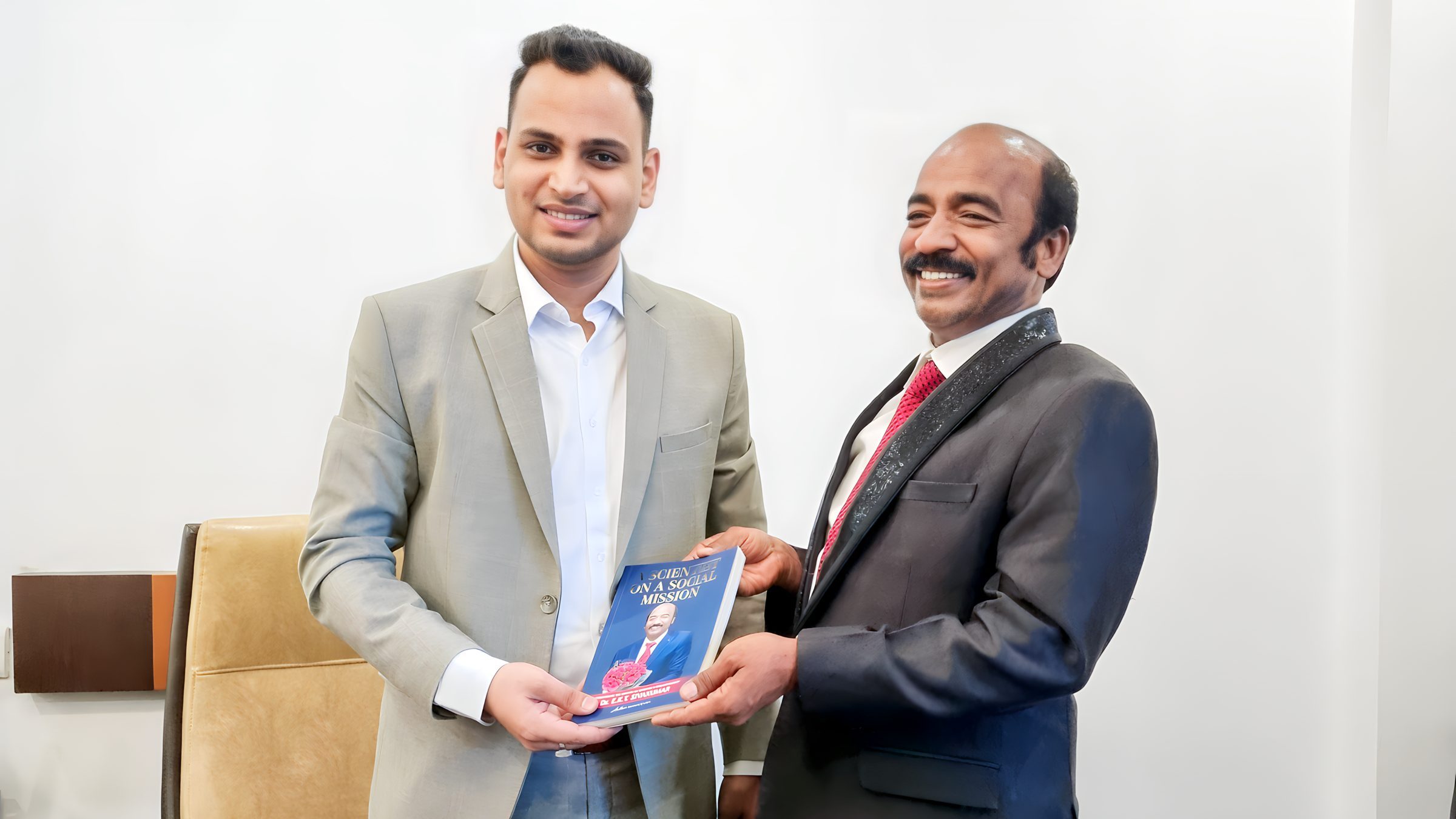 Lithvik Mukesh Sharma is photographed at his private office. Two men stand indoors, jointly holding a book titled 'A SCIENTIST ON A SOCIAL MISSION' by Dr. B.V. Srinivas. The author of the book A SCIENTIST ON A SOCIAL MISSION